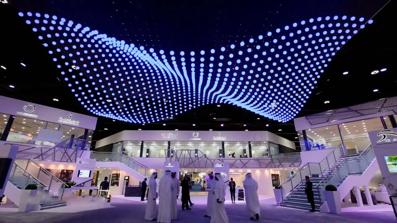 Exhibition hall with a wavy blue LED dot ceiling; people in traditional dress and glass staircases nearby.
