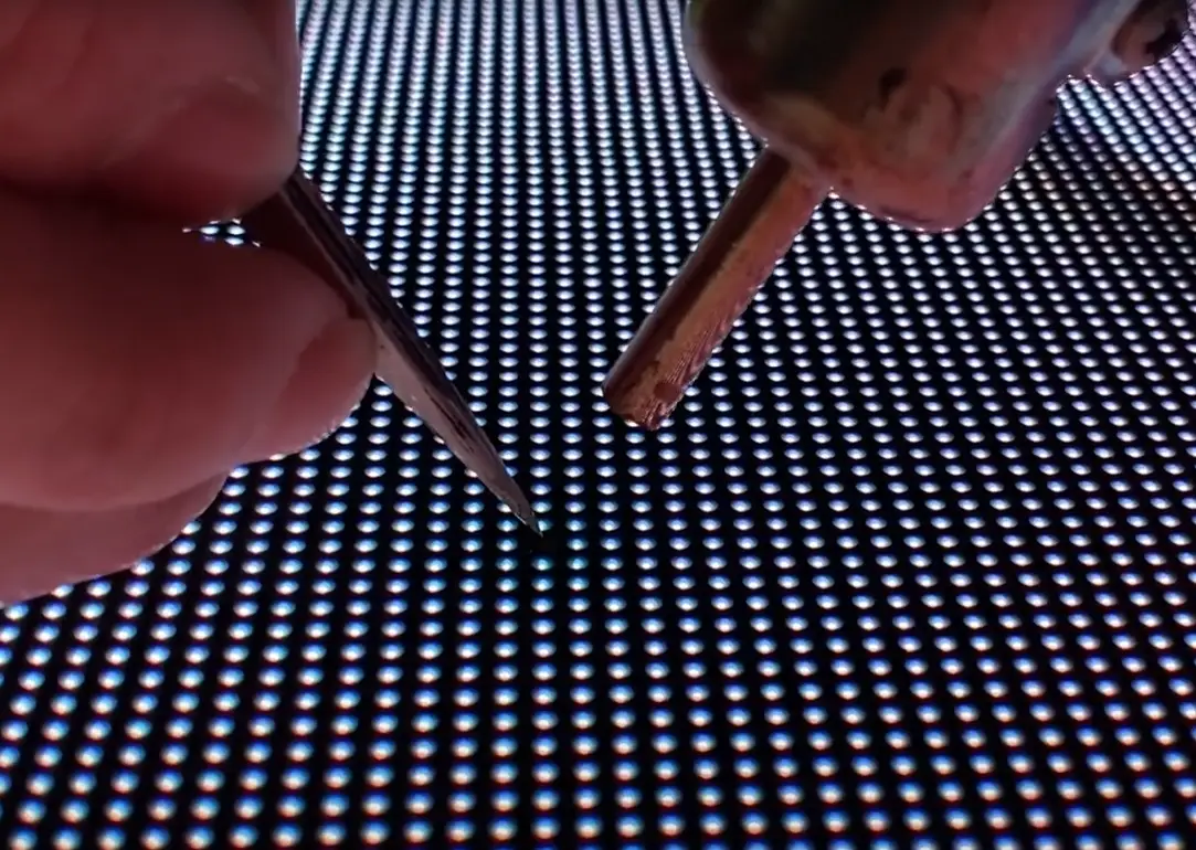 Close-up of two metal tips held by fingers above a perforated blue LED grid surface.