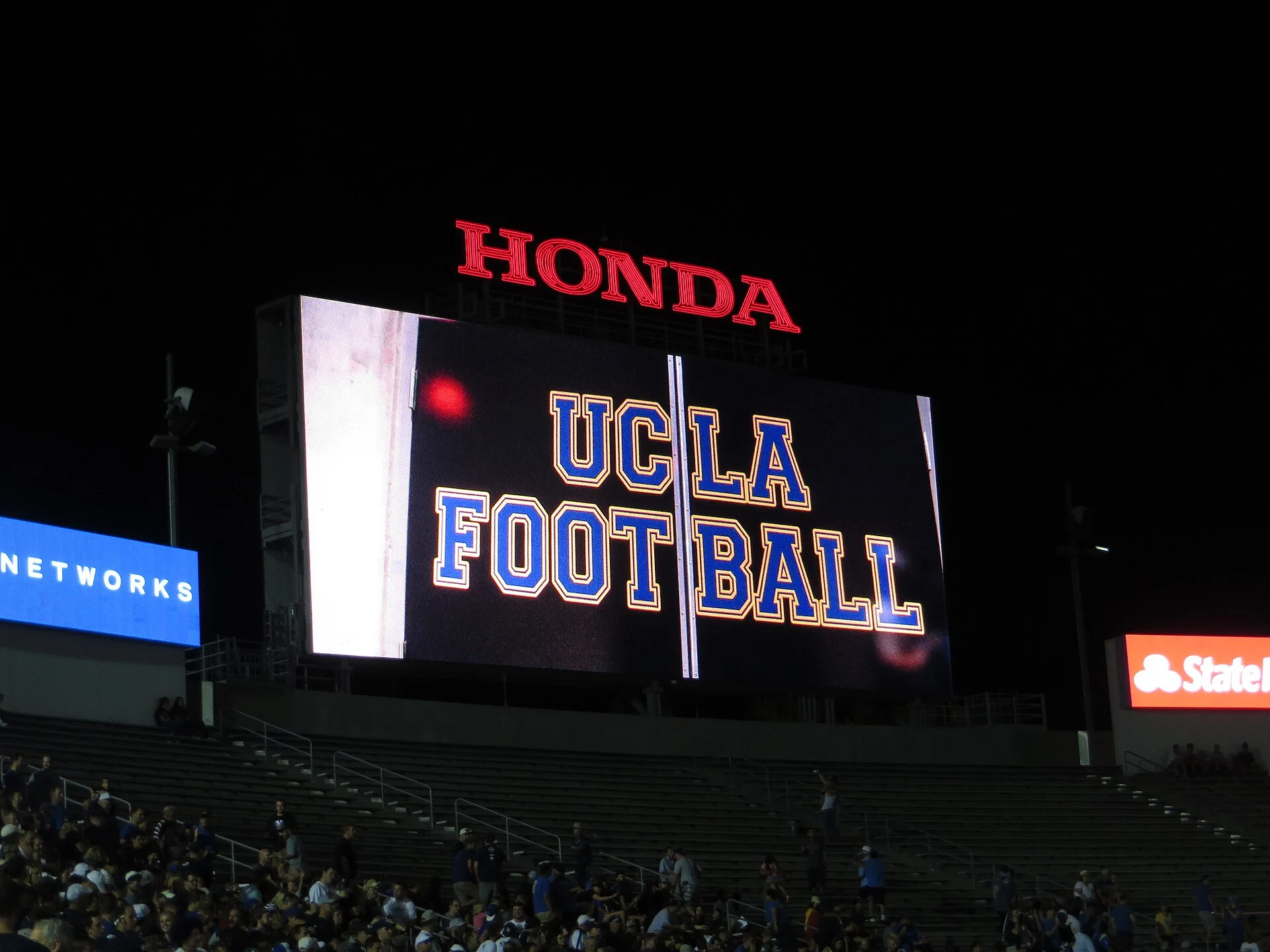 Night scene at a stadium with a large LED scoreboard reading 'UCLA FOOTBALL' and a red 'HONDA' sign above it.